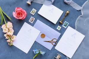 Wedding stationery suite with white invitations, pink roses, and vintage stamps arranged on blue fabric background.