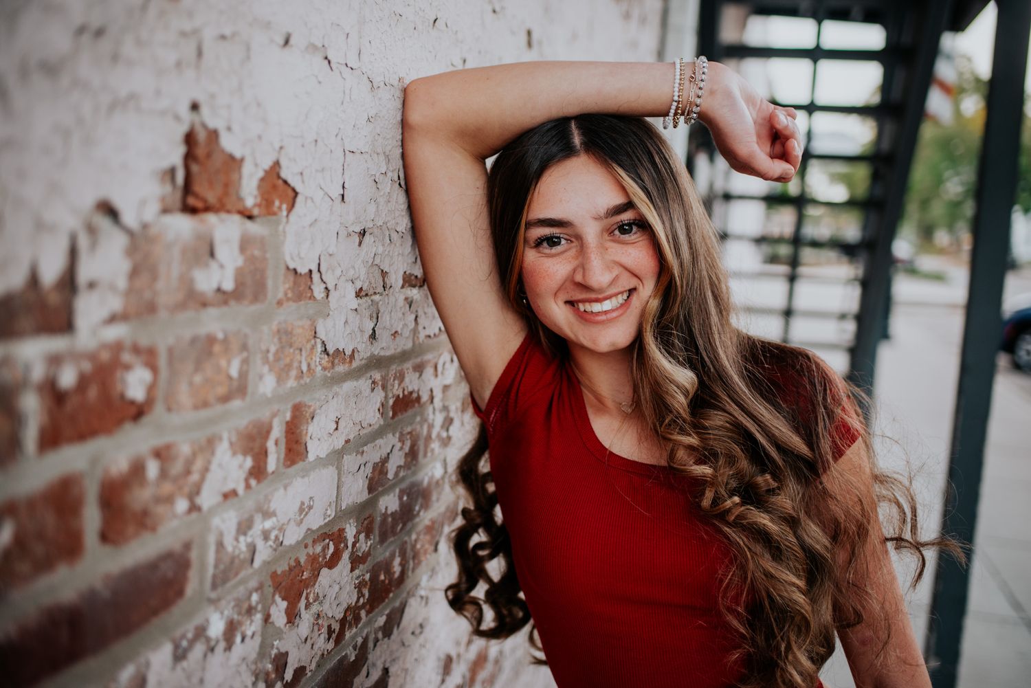 High schooler in a red sweater poses against a rustic brick wall with natural lighting during birthday photo shoot.