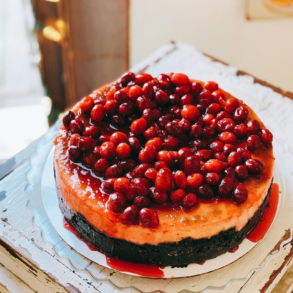 A decadent cheesecake topped with fresh cranberries and dark chocolate cookie crust on a white rustic surface.