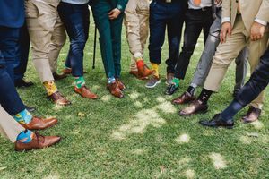 Wedding party members show off their colorful socks while standing in a line on grass.