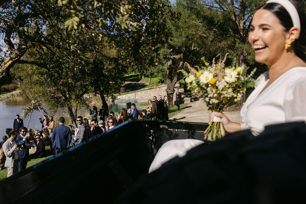 A joyful bride in a white dress holds a bouquet while guests gather at an outdoor wedding ceremony by a lake.
