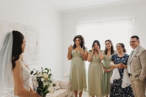 A wedding party gathers in a bright white room with floral arrangements doing a first look with the bride