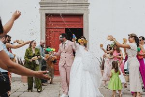 Wedding guests toss flower petals and celebrate as newlyweds exit a white church building with red doors.
