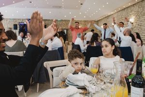 Guests raise their hands in celebration at a wedding reception while two young children sit at a dining table.