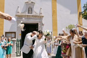 Newlyweds exit ornate white church doorway while guests shower them with flower petals in joyful celebration in LIsbon