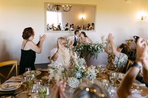 Wedding dinner scene with guests raising hands in joy at a rustic table decorated with white floral centerpieces.