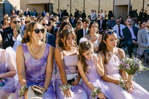 Bridesmaids in lavender dresses sit together outdoors at a sunny wedding ceremony holding delicate bouquets in Lisbon