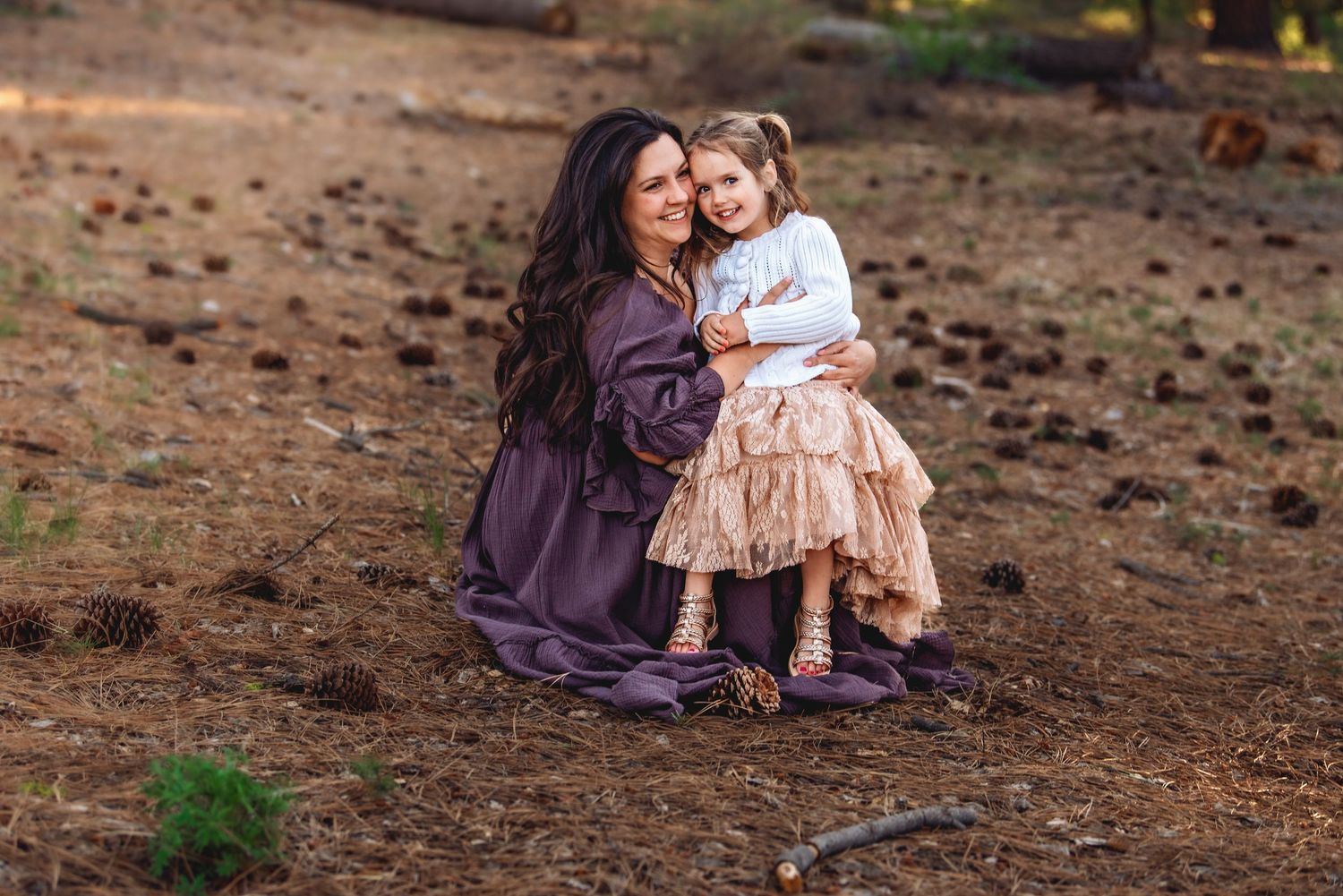A mother and daughter share tender moments together while sitting on brown soil during an outdoor photo session.