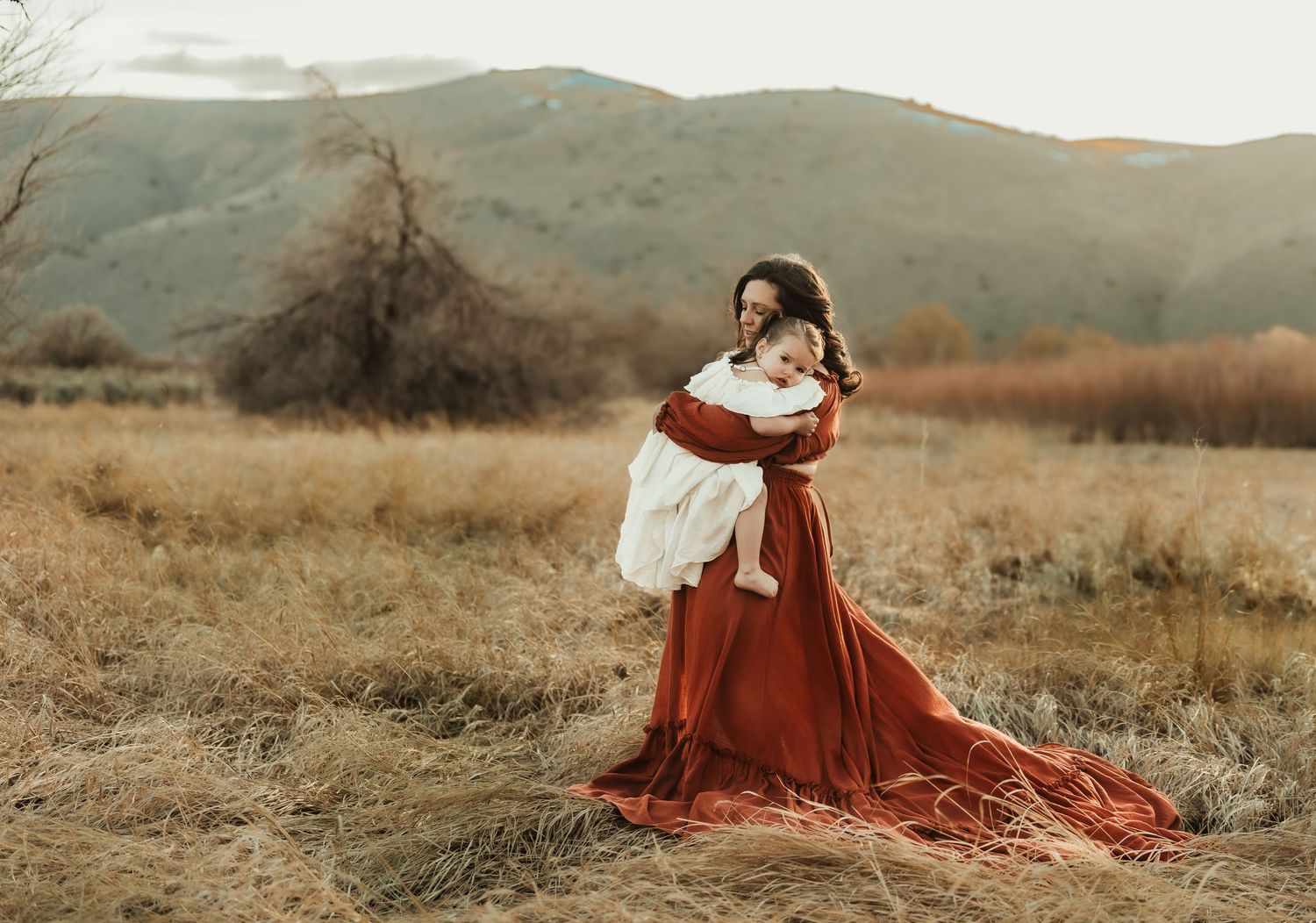 Dramatic portrait in flowing red dress holding baby against mountain backdrop.