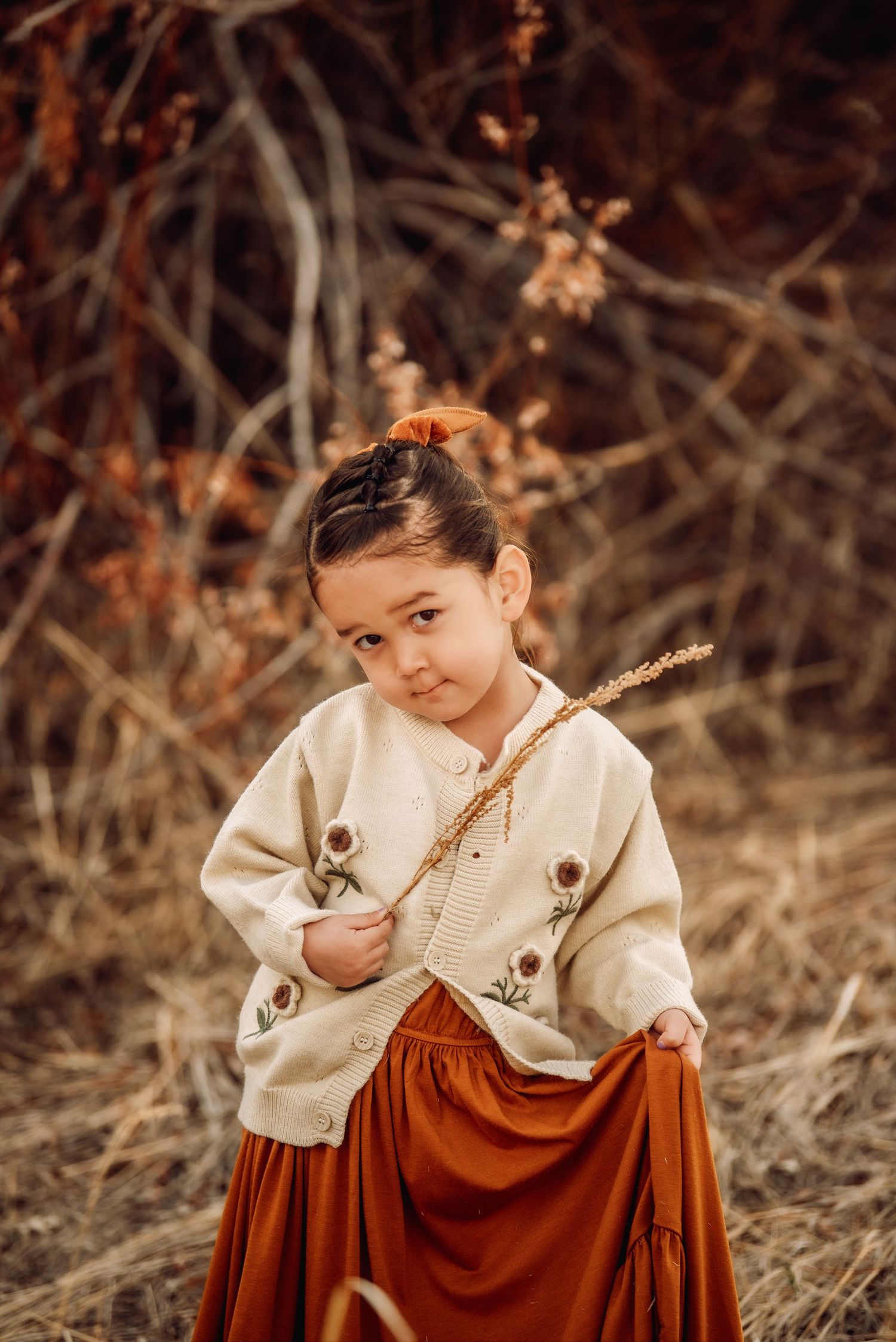 A young child in a vintage-style cream blouse and brown dress holds a small twig while sitting outdoors in autumn.