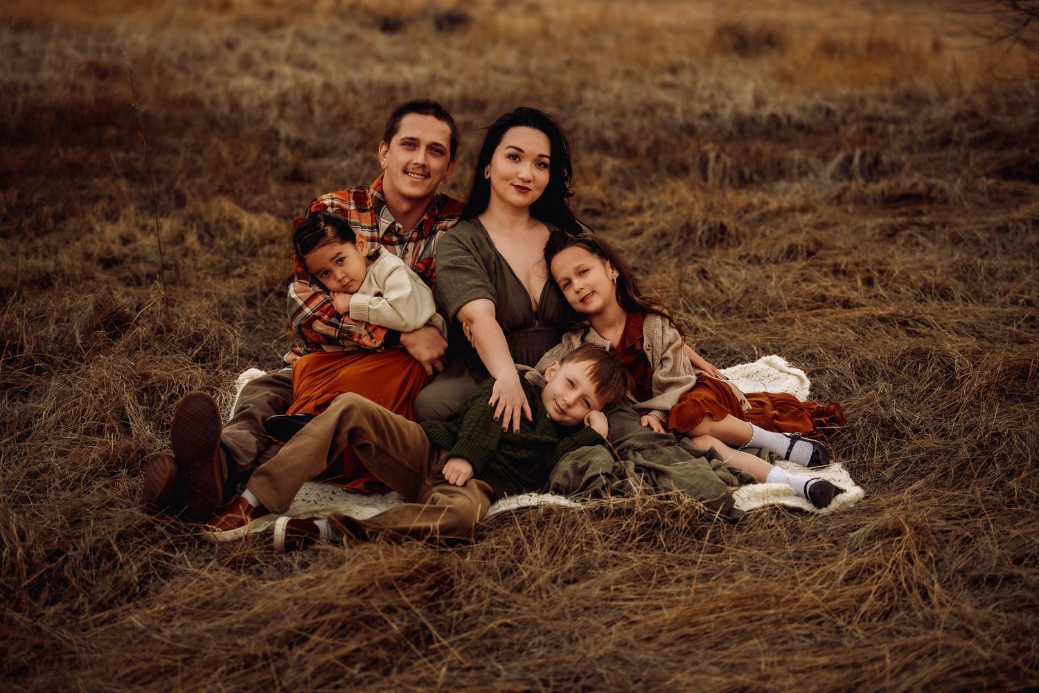 A couple sits together on brown grass during an outdoor autumn photoshoot in warm earth tones.