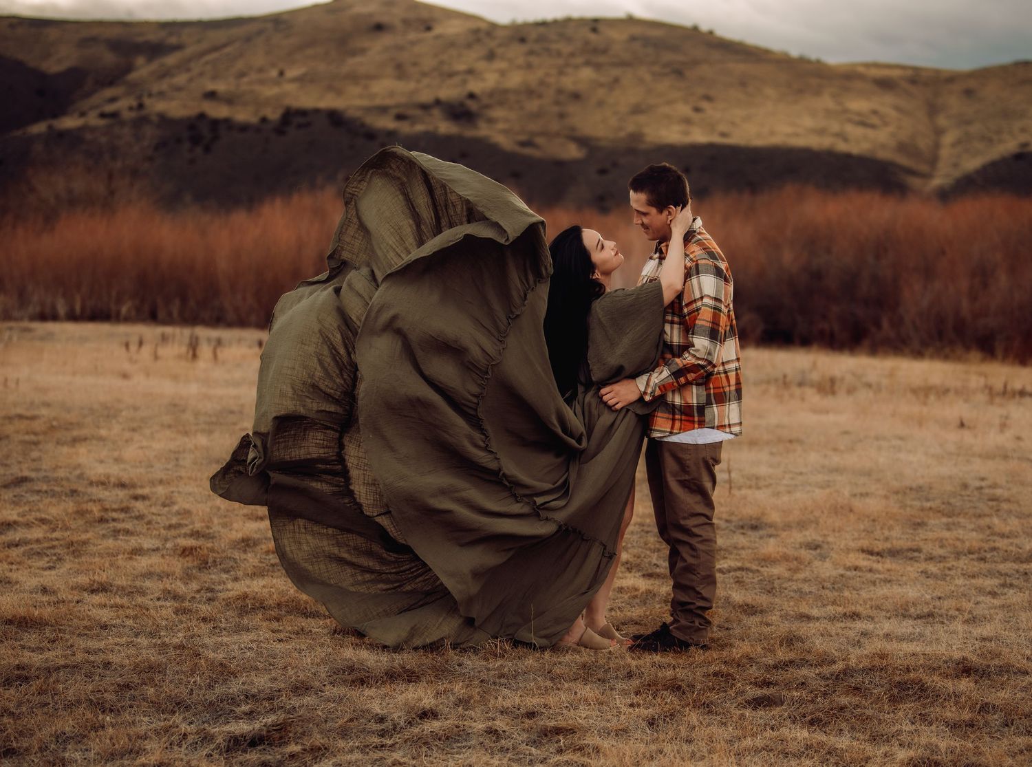 Two people in warm outdoor clothing embrace while sitting in a rustic autumn field at sunset.
