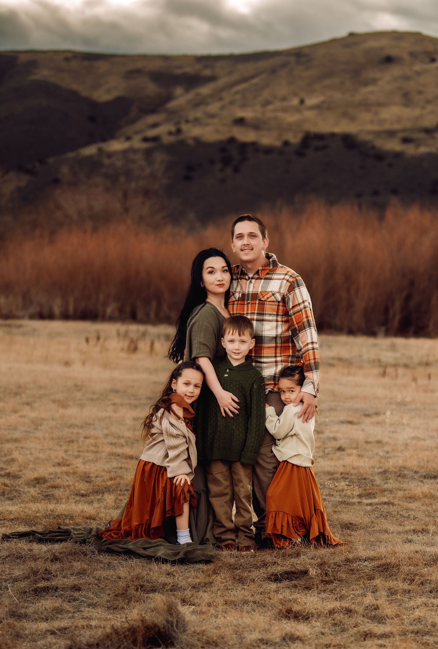 A family sits together in a rustic field with brown grass and rolling hills in the background during golden hour.