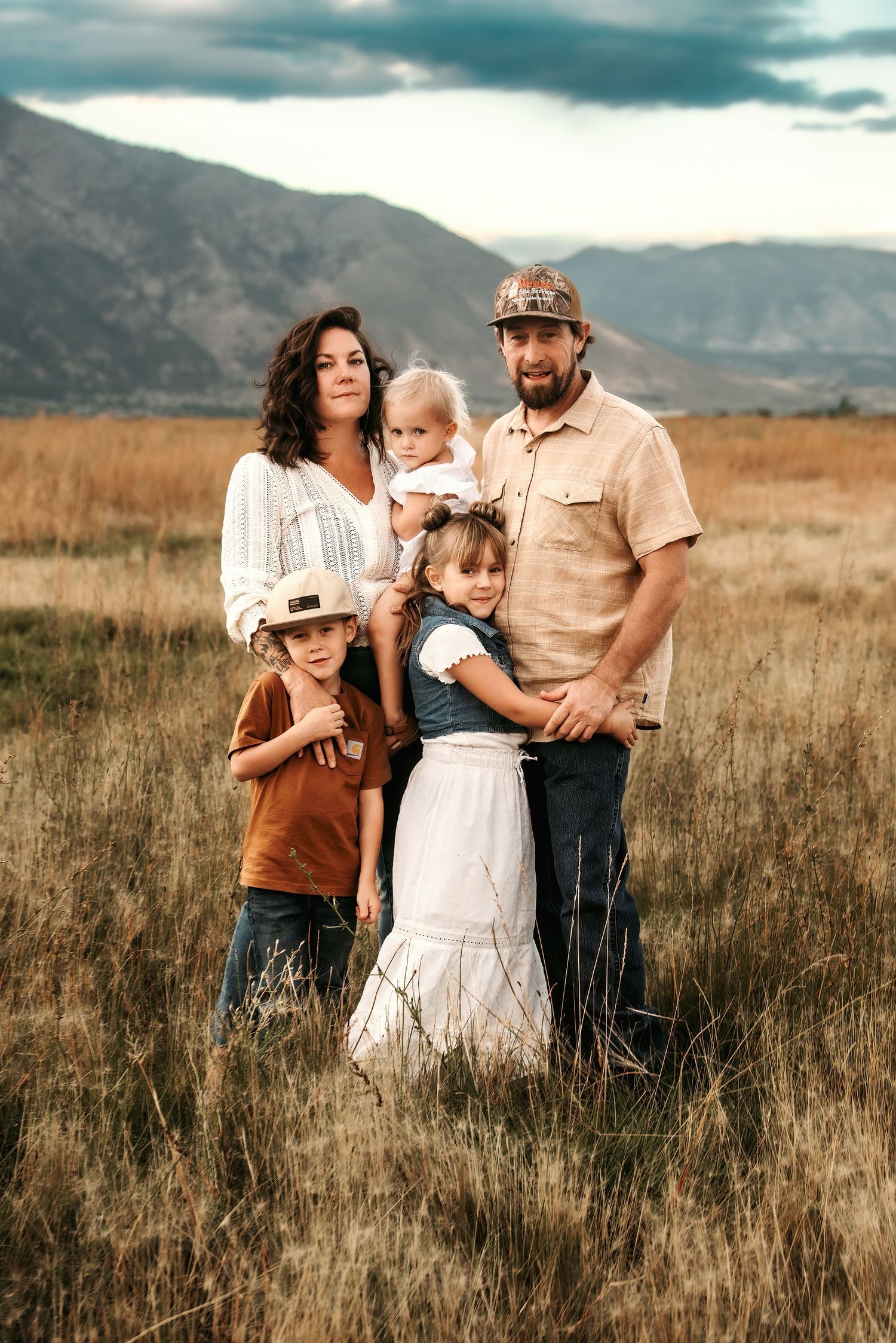 Family in western attire embraces together in golden field during sunset with mountains in background.