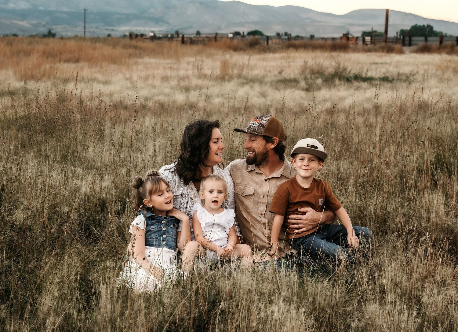 A family sits together in a golden field of tall grass with mountains visible in the distance at sunset.