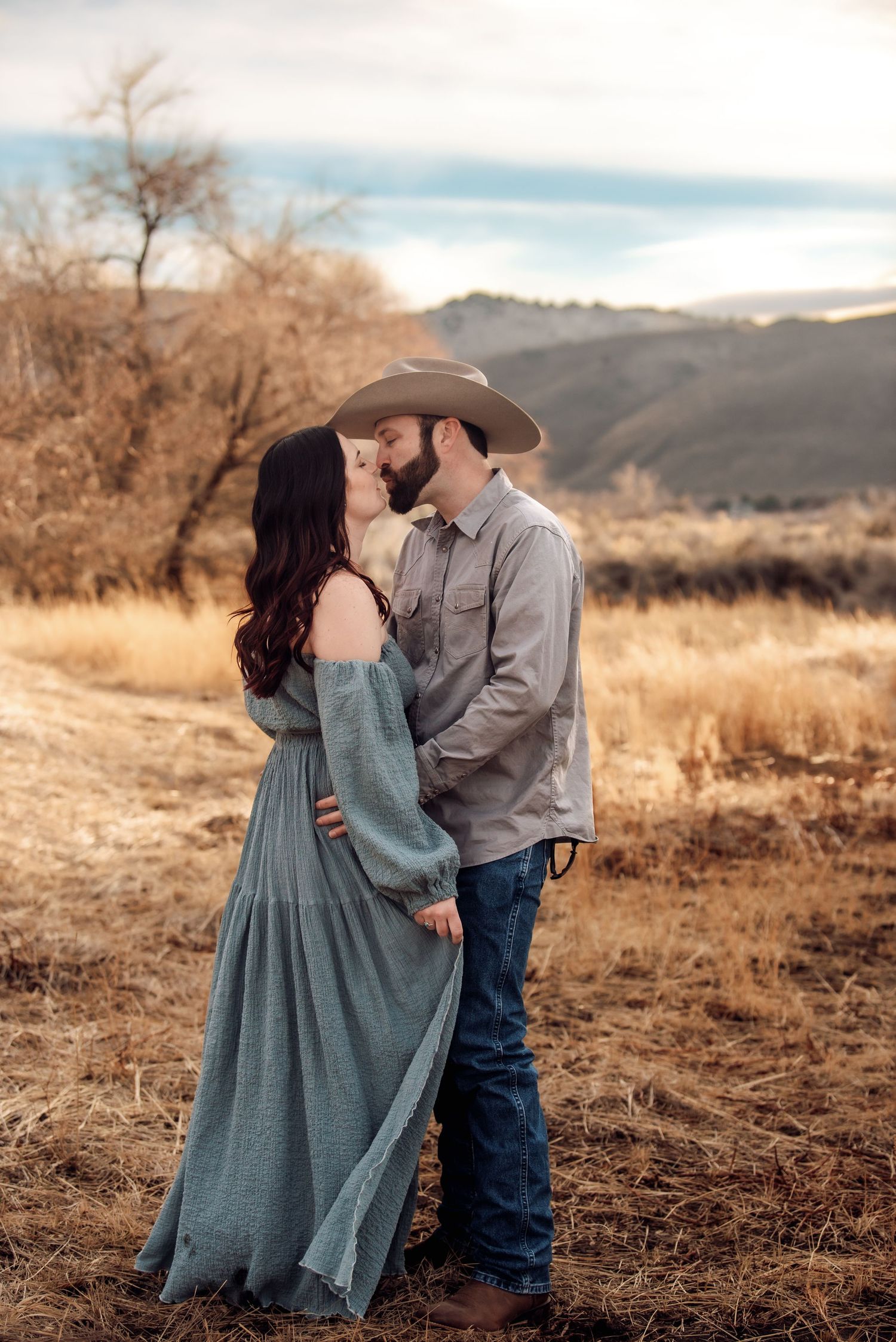 A couple shares a romantic moment in a rustic field during golden hour with mountains in the background.