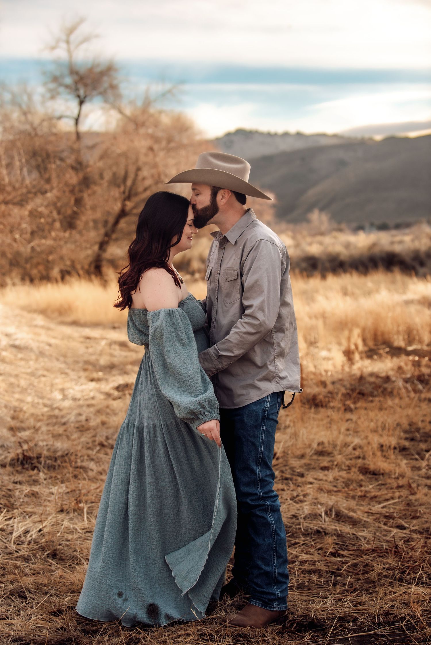 A romantic couple in western attire embrace on a hillside during golden hour against a scenic mountain backdrop.
