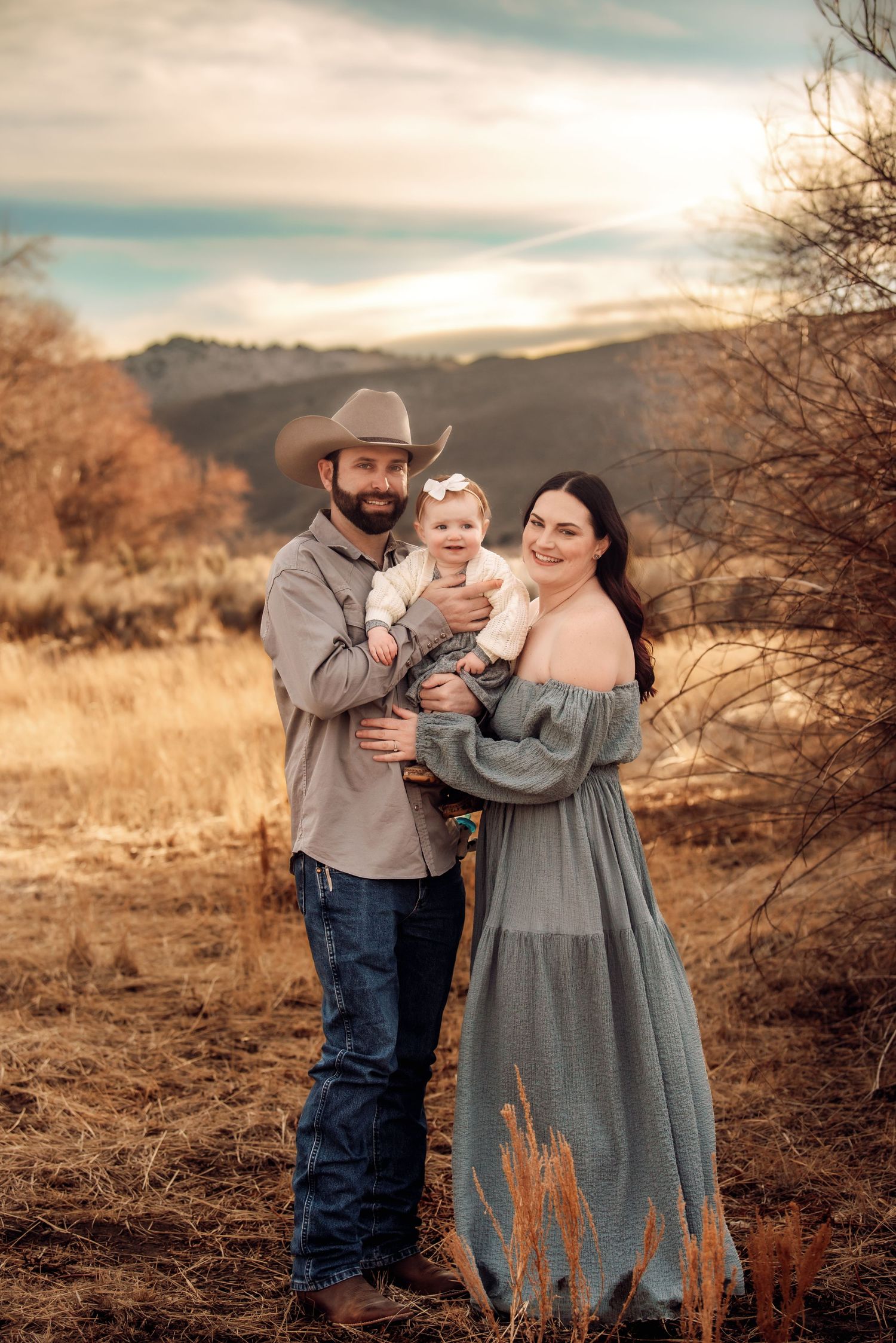 A family in western attire poses together in a golden field during sunset with mountains in the background.