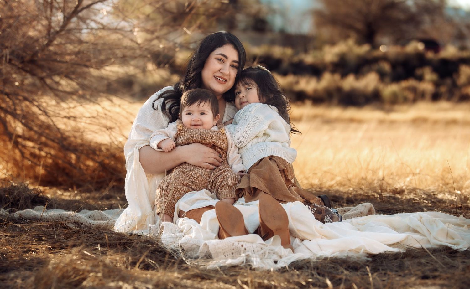 A family wearing white outfits embraces while sitting on a blanket in a golden field at sunset.