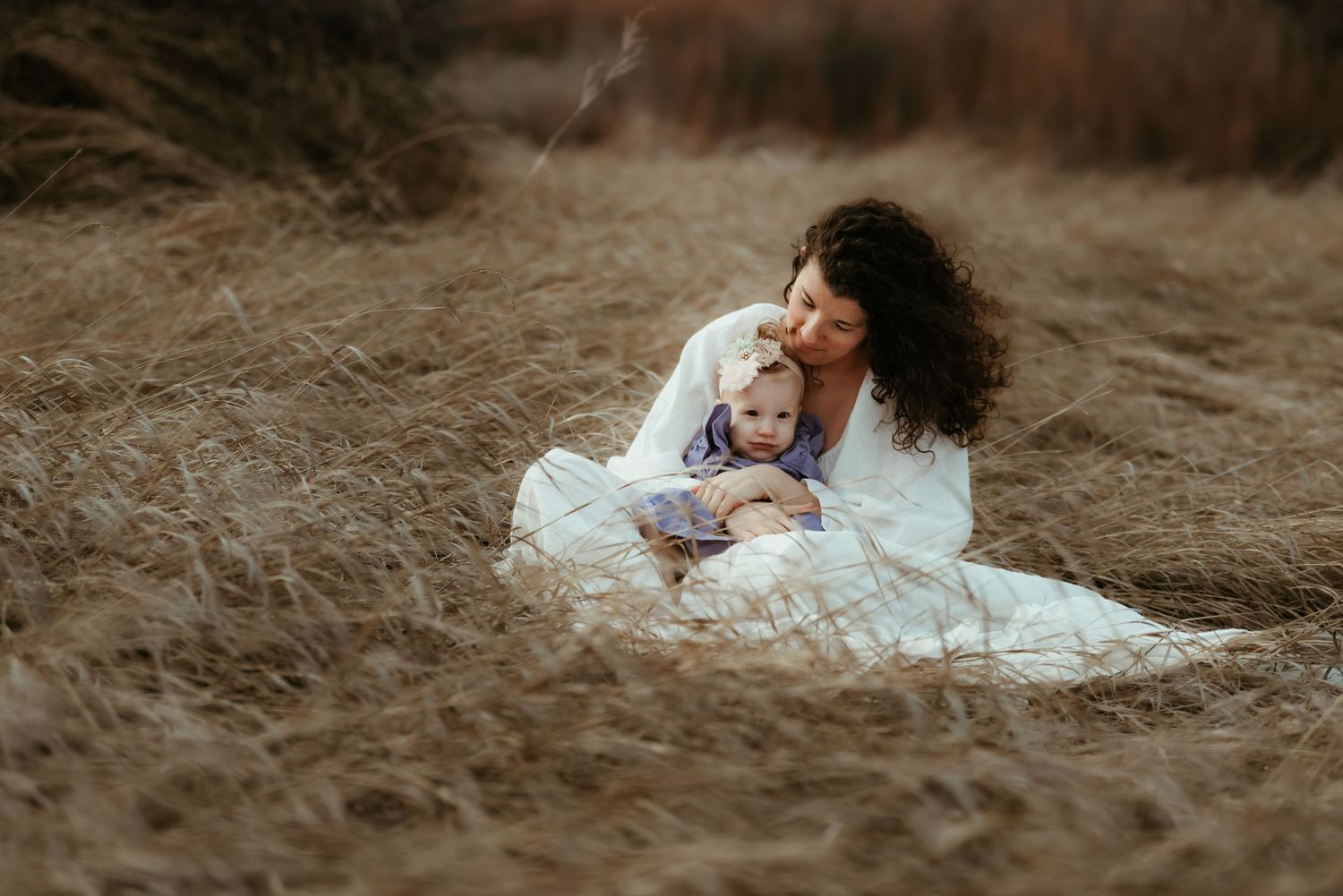 Intimate outdoor portrait with figure in white dress holding baby in field.