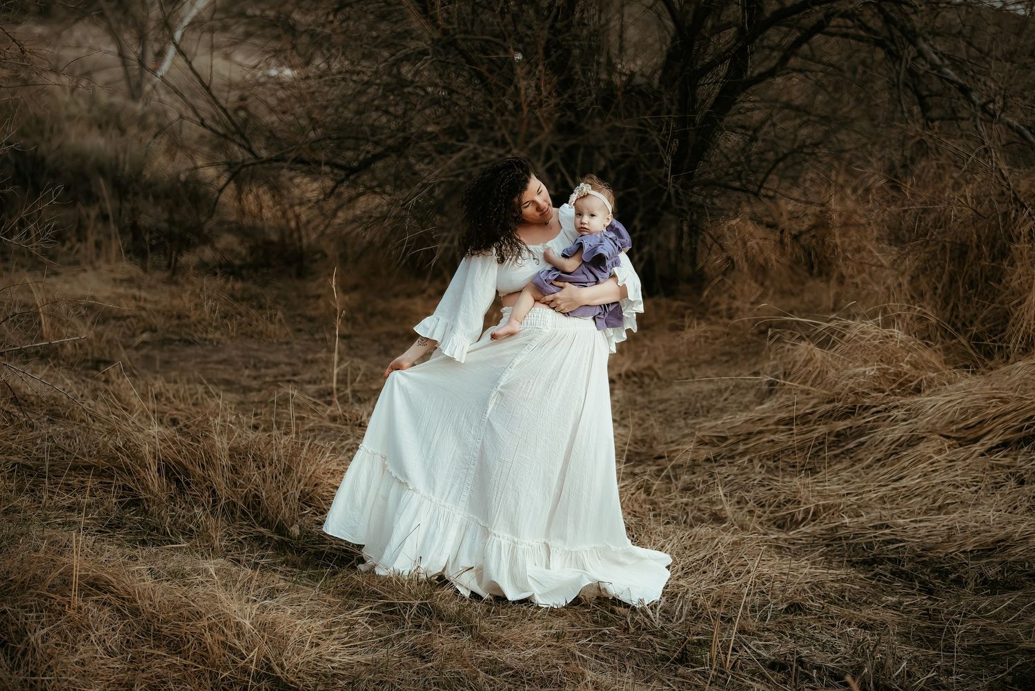 Figure in flowing white dress holds baby in rustic outdoor setting.