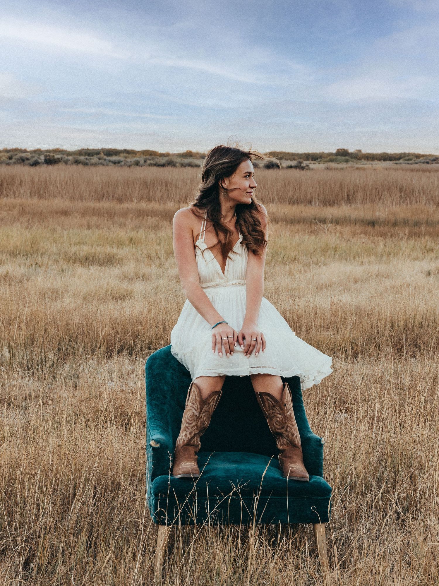 Person in a white dress and cowboy boots poses on a vintage chair in a rustic field.
