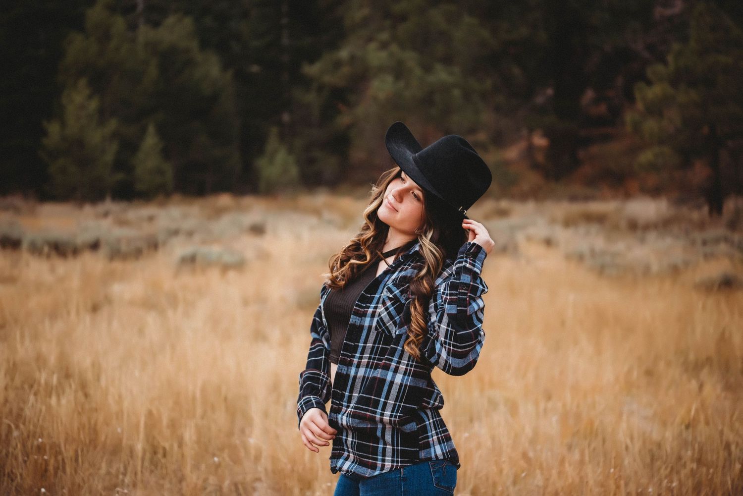 Person in plaid shirt and hat poses against autumn field and forest backdrop.