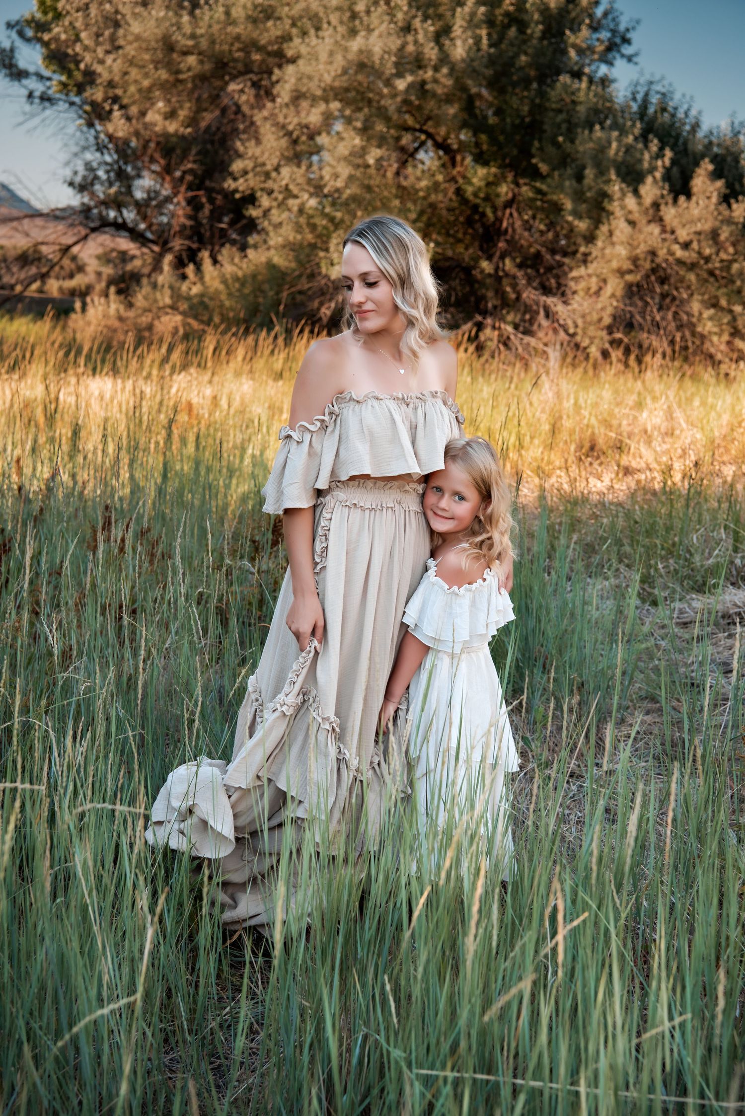 Mother and daughter in matching off-shoulder dresses stand in tall grass at sunset.
