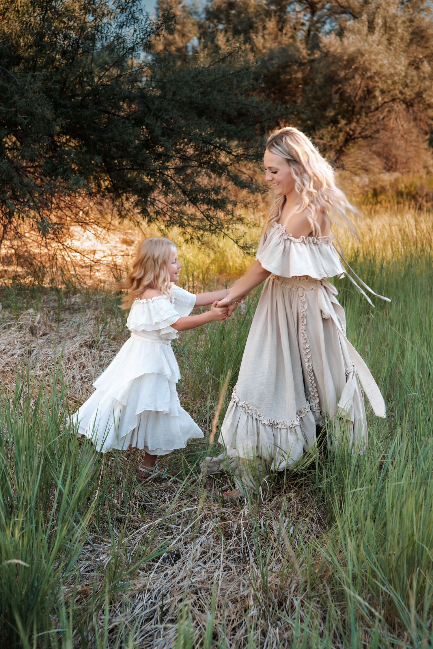 Mother and daughter in matching white dresses dance in sunlit meadow.