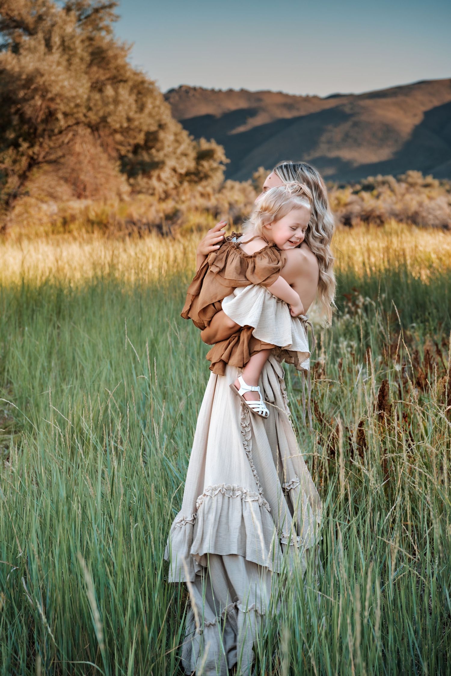 Woman in flowing white dress holds child in brown outfit among tall grasses.