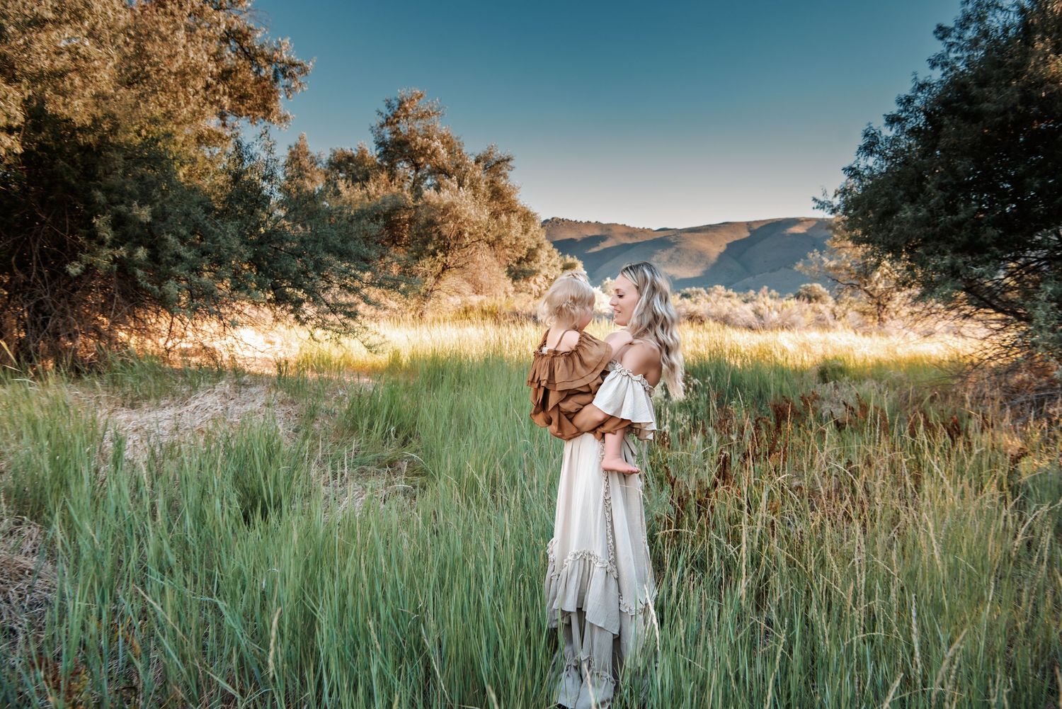 Woman in white dress holds child in sunlit meadow with mountains in background.