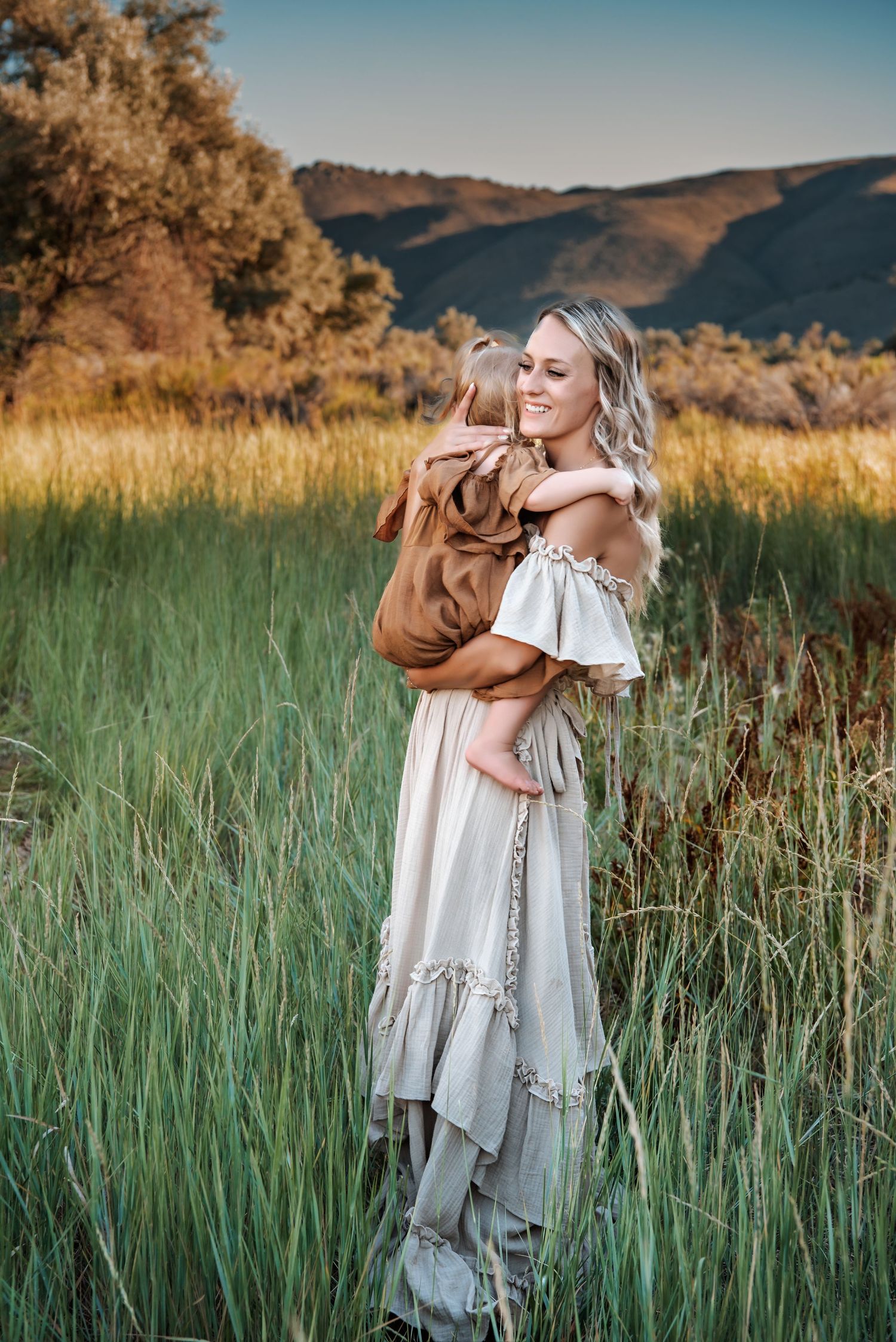 Woman in off-shoulder dress holds child against mountain backdrop.