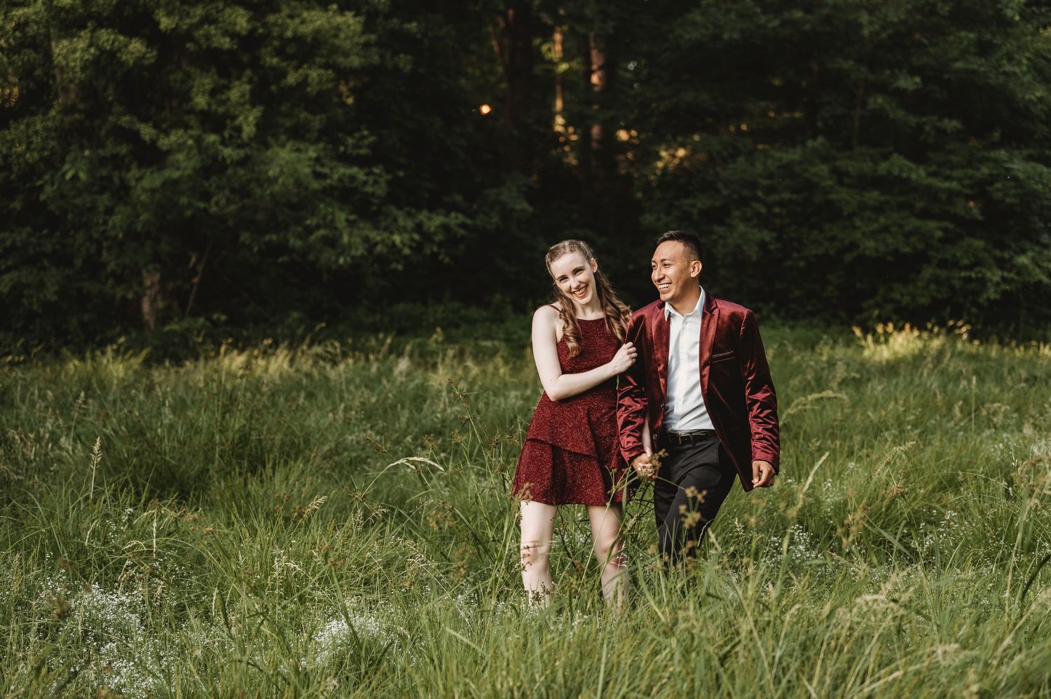 A couple in burgundy attire shares a moment while walking through a grassy field at dusk.