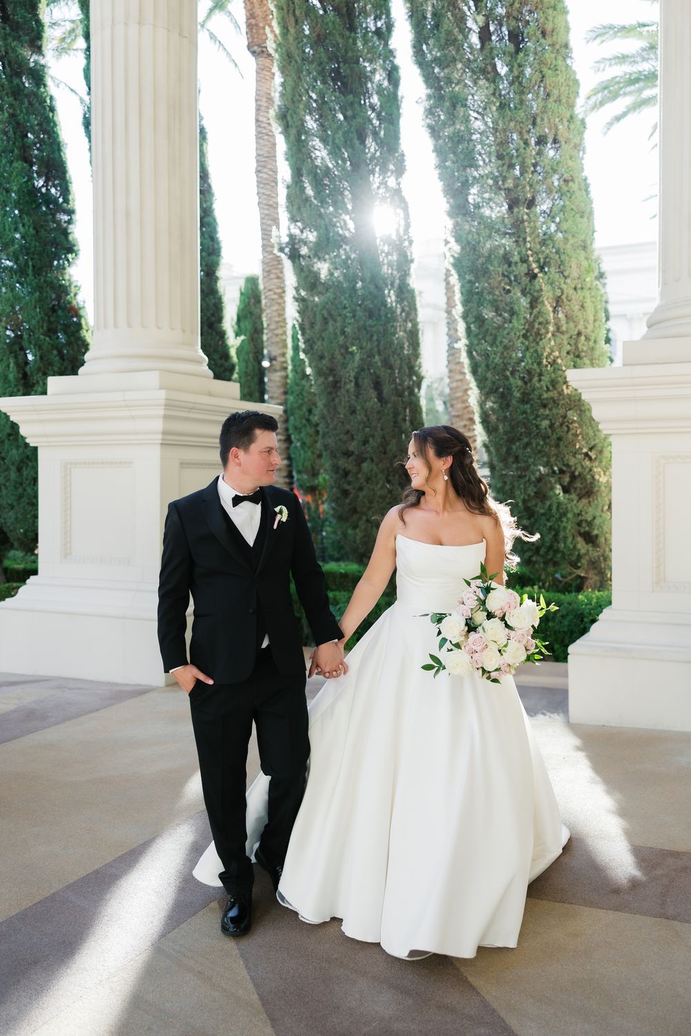 A bride in a white ballgown and groom in black tuxedo walk hand-in-hand between elegant white columns and cypress trees.