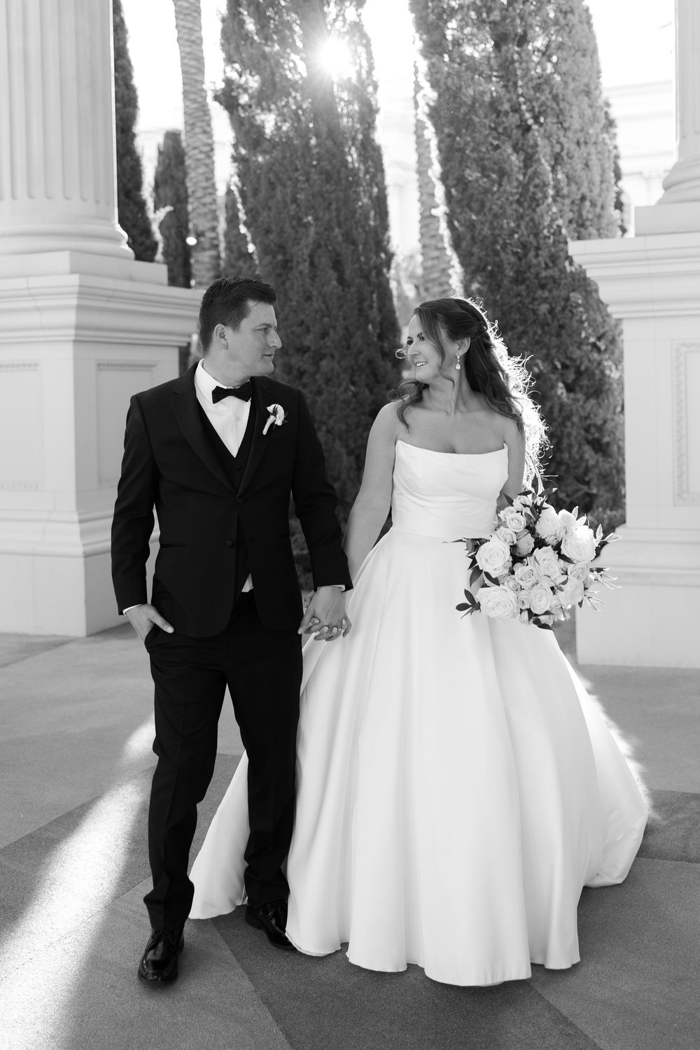 A bride and groom walk together through an elegant colonnade with white pillars and cypress trees in the background.