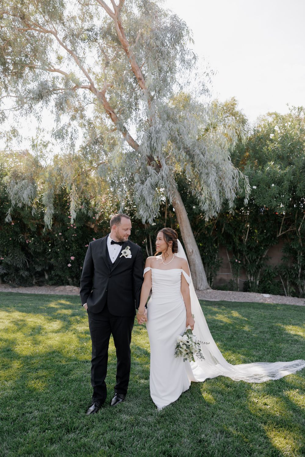 A bride and groom share a romantic moment together on their wedding day in a sunlit garden with willow trees.