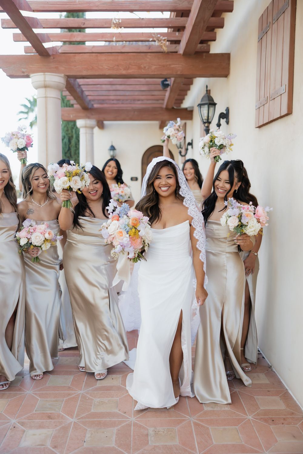 Bridal party in champagne colored dresses dancing and celebrating in a Spanish-style hallway with terracotta floors.
