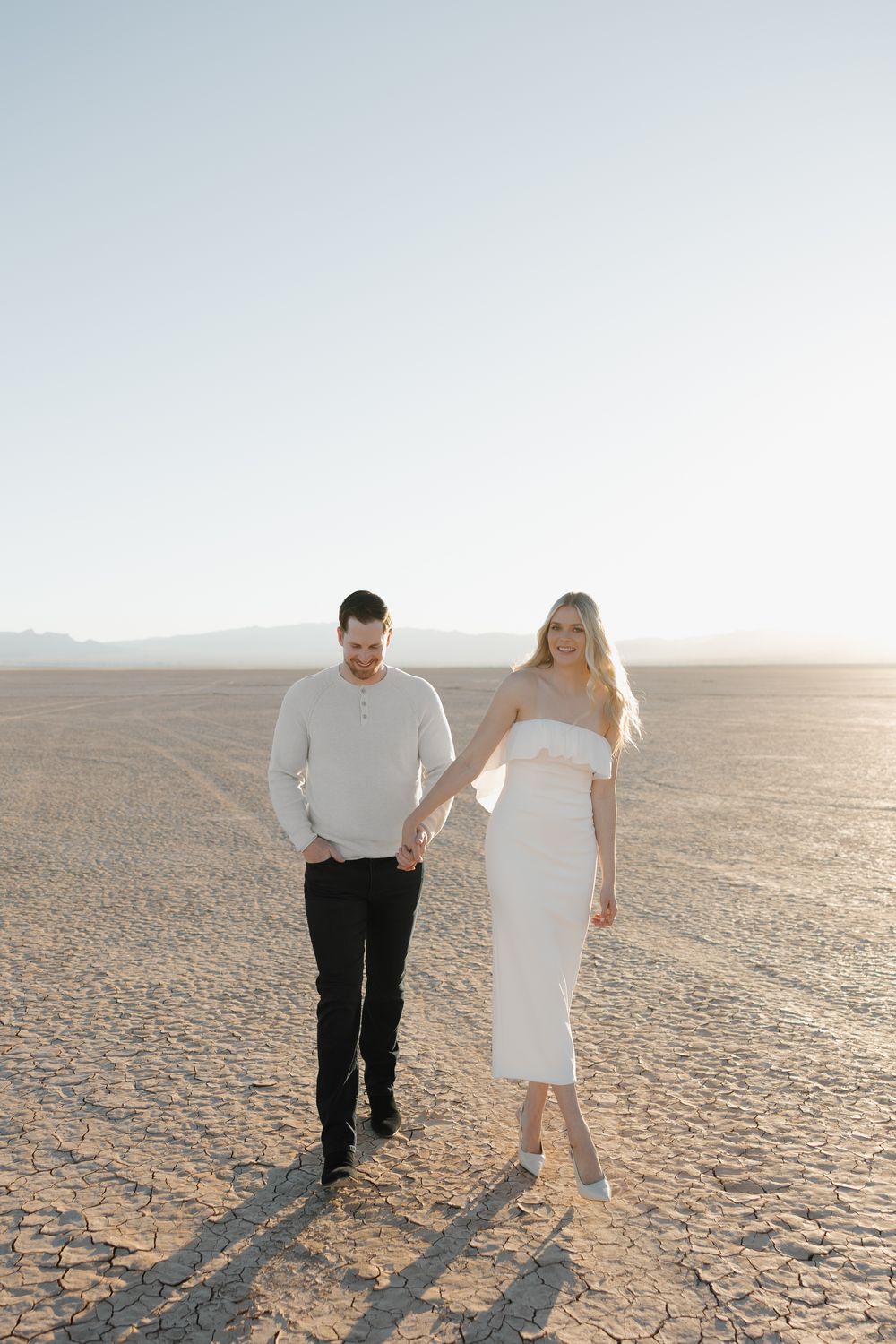 A couple stands together on a vast dry desert landscape at sunset casting long shadows on the cracked earth.