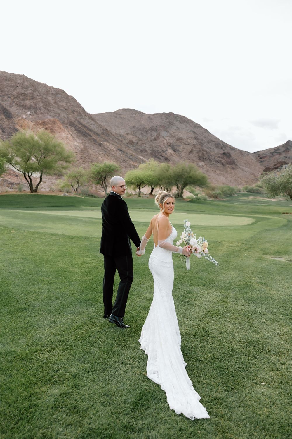 A couple in wedding attire stands on a lush golf course with rugged desert mountains in the background.