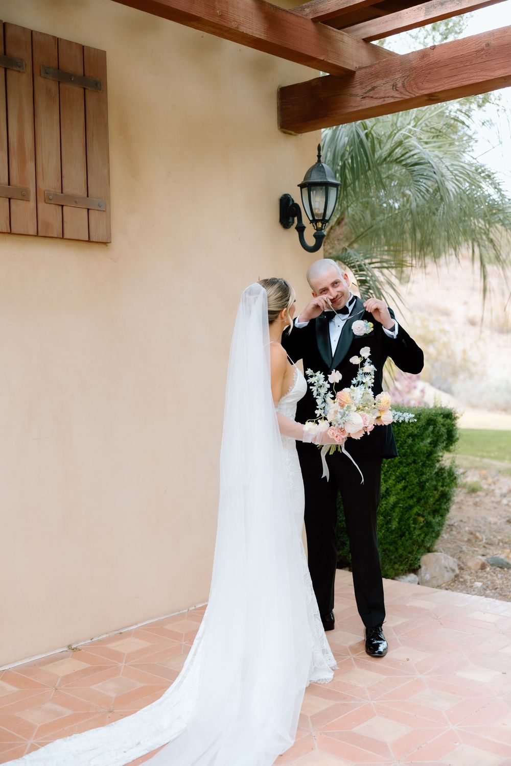 A romantic first look moment between a bride in white gown and groom in black tuxedo on a tiled patio.