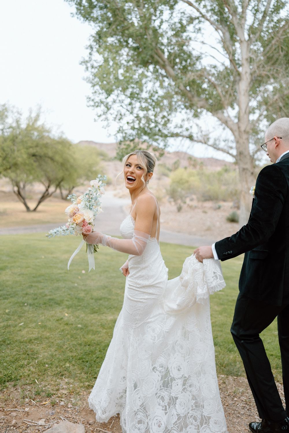 A couple in wedding attire playfully blows bubbles together on a grassy hill with trees in the background.