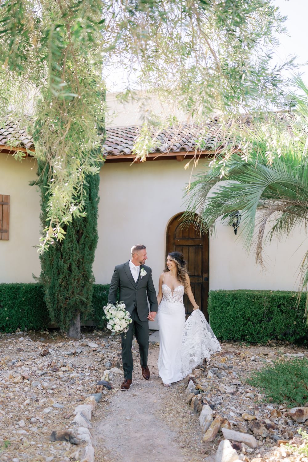 A couple in formal attire walks along a garden path with palm trees and greenery at an outdoor venue.