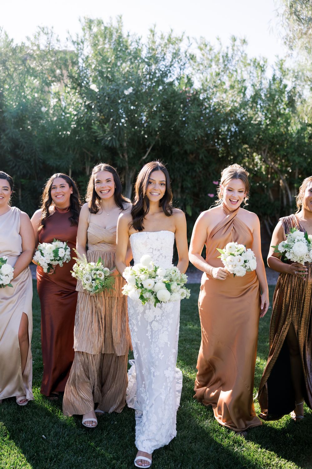 A group of bridesmaids in earth-toned dresses stand together outdoors holding white bouquets in sunlit garden setting.
