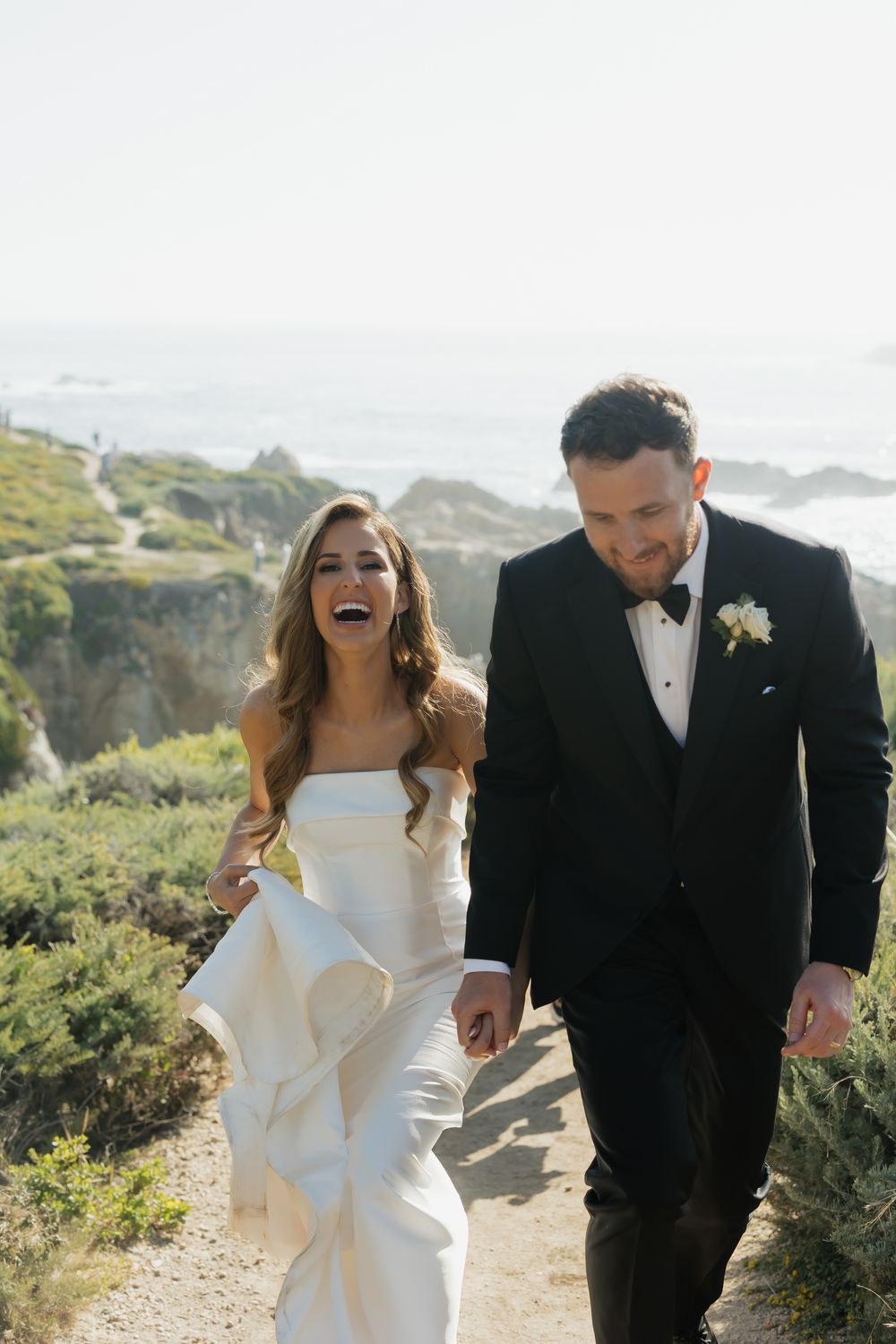 Joyful wedding couple walking and laughing together on coastal cliffs with ocean views.