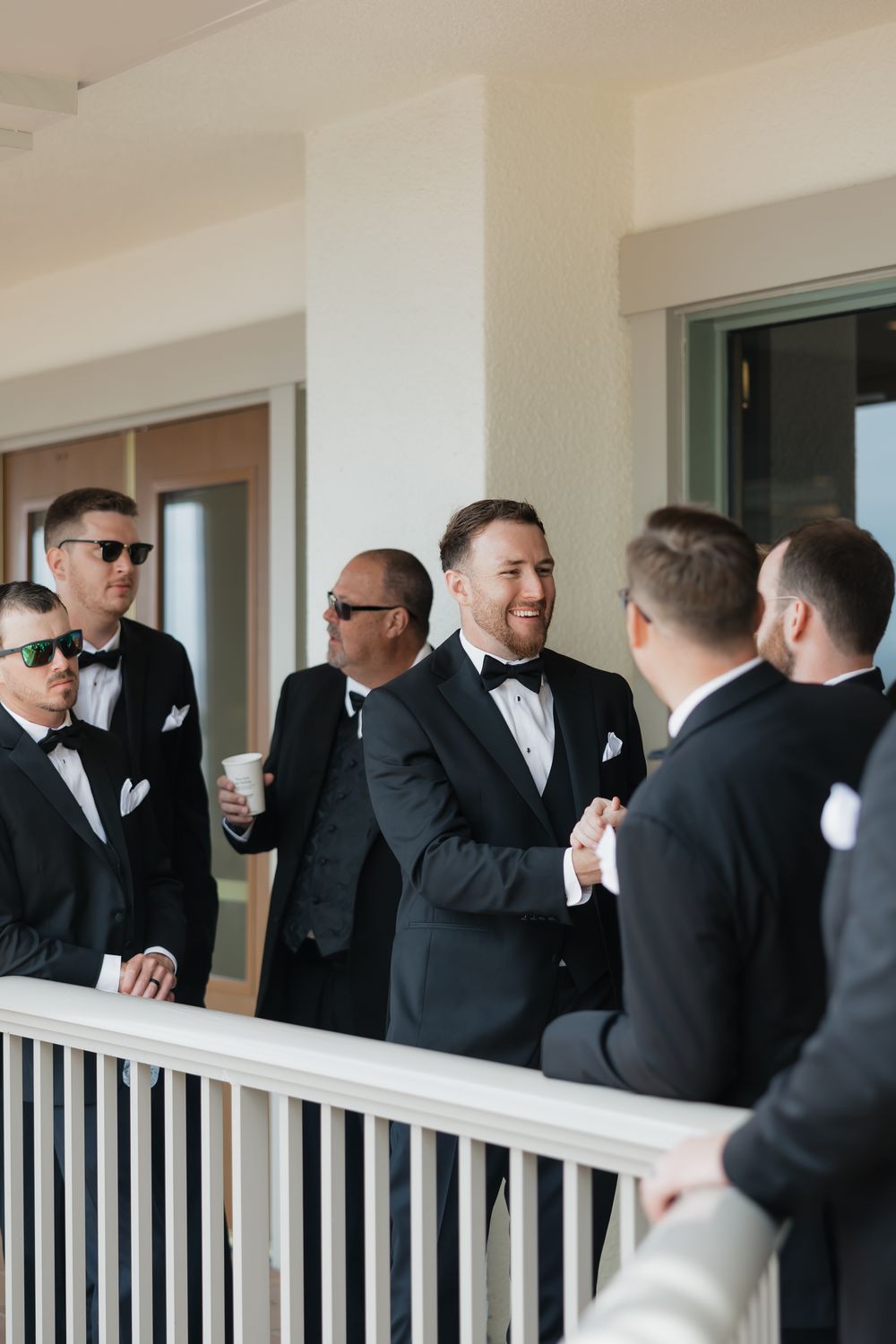 Groomsmen in black tuxedos and sunglasses gather on a white balcony sharing laughs and drinks before the wedding.