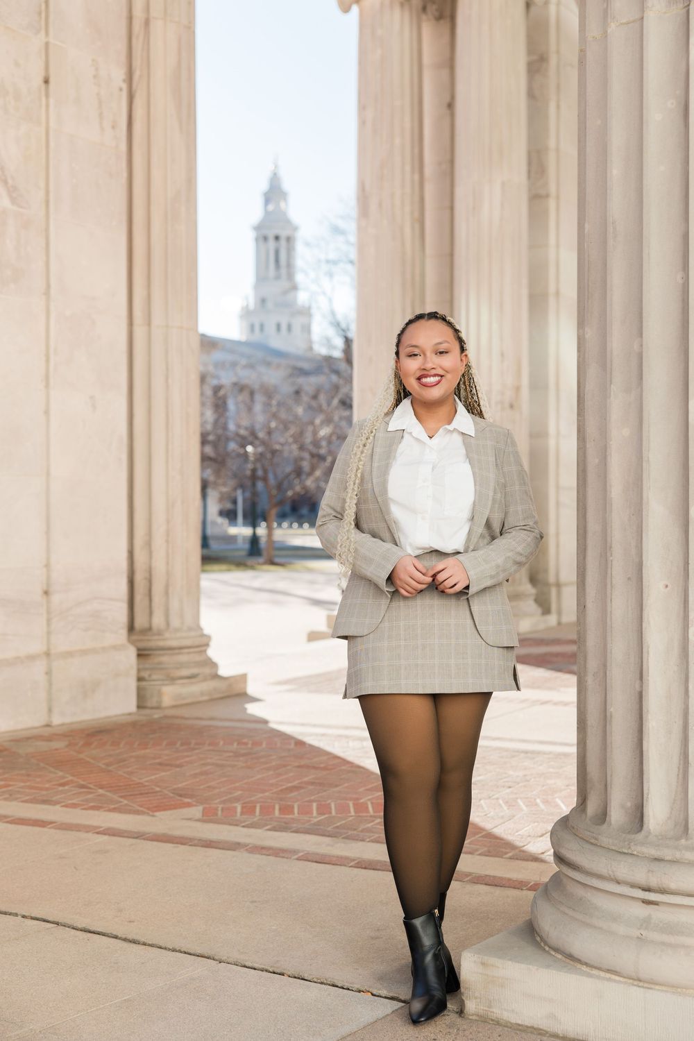 A student poses between classical columns on a college campus with a historic building and bell tower in the background.