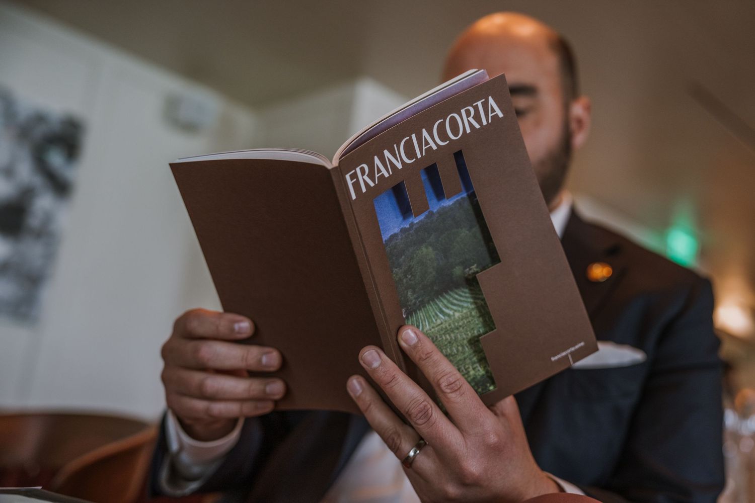 A person holds a brown hardcover book in a dimly lit room, with multiple similar books visible in the background.