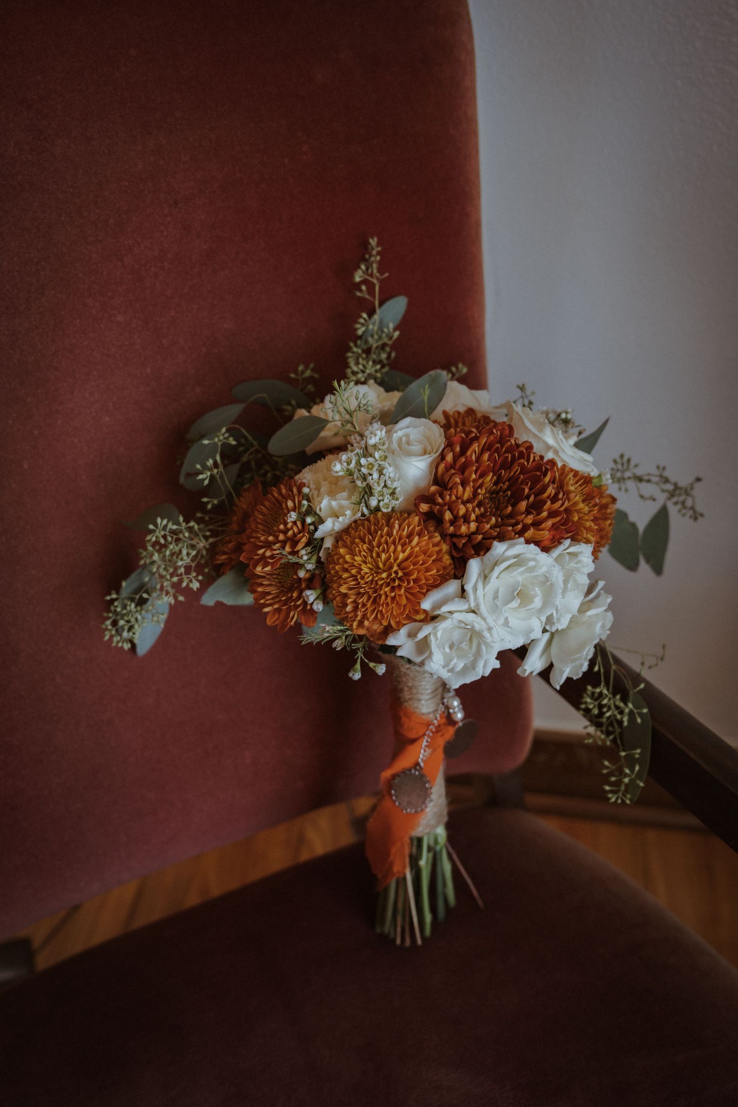Wedding bouquet with white roses and orange chrysanthemums against a burgundy background in moody lighting.