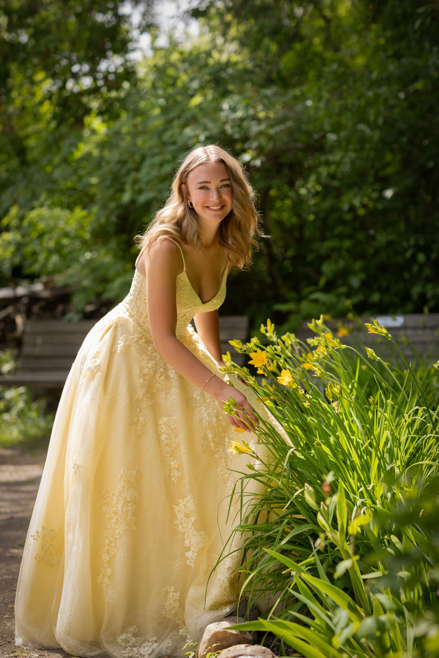 A person in a flowing yellow formal dress bends down to admire blooming daylilies in a garden setting.