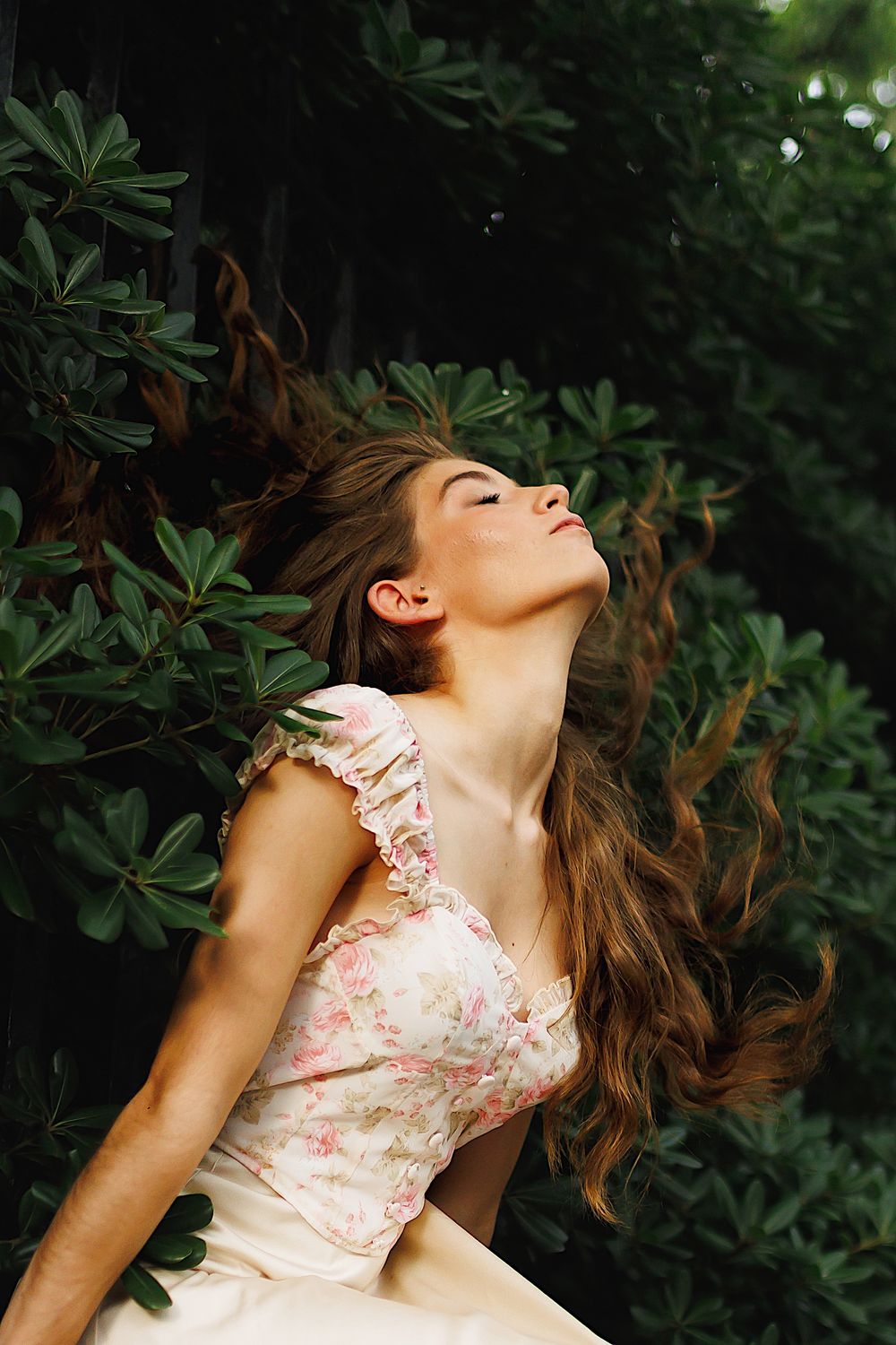 Flowing hair catches sunlight against green foliage backdrop as figure in pink floral dress leans back in dreamy pose.